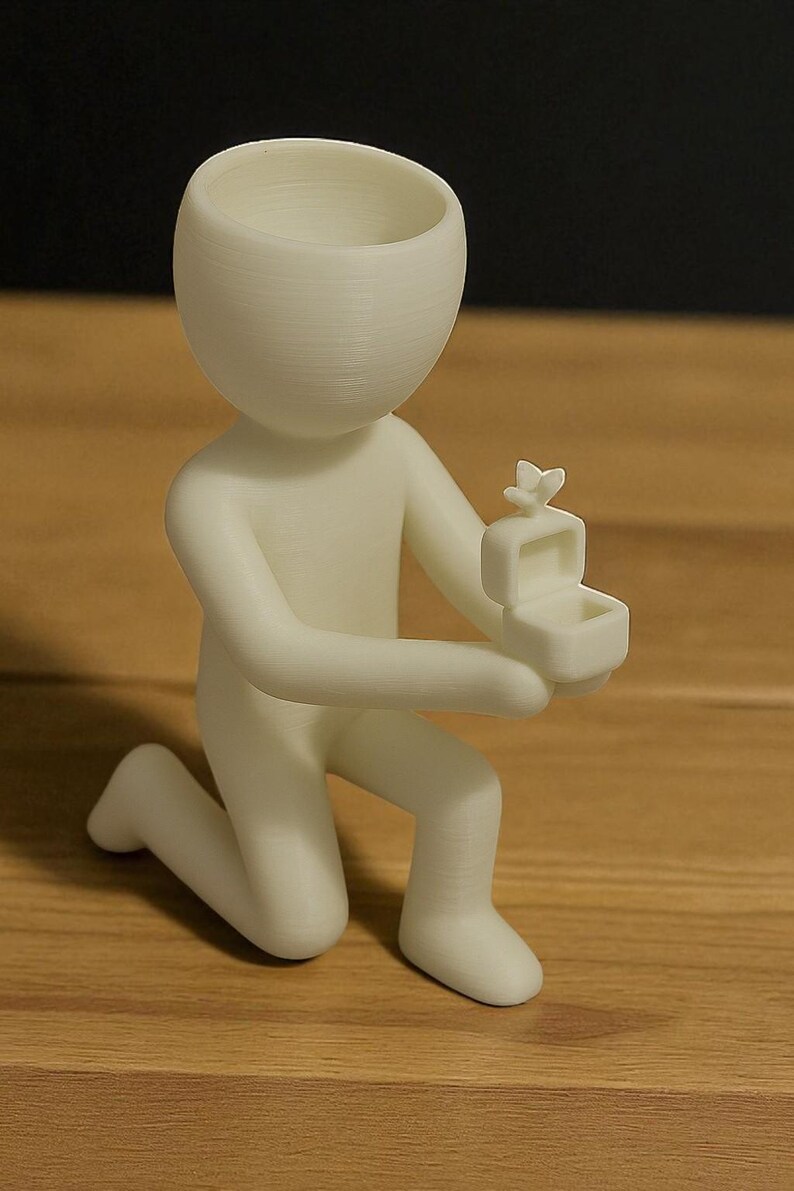 May include: A white figurine of a person kneeling, holding a small box with a butterfly on top. The figurine has a large, open head and is positioned on a wooden surface. The box is open, suggesting a ring or small item inside.
