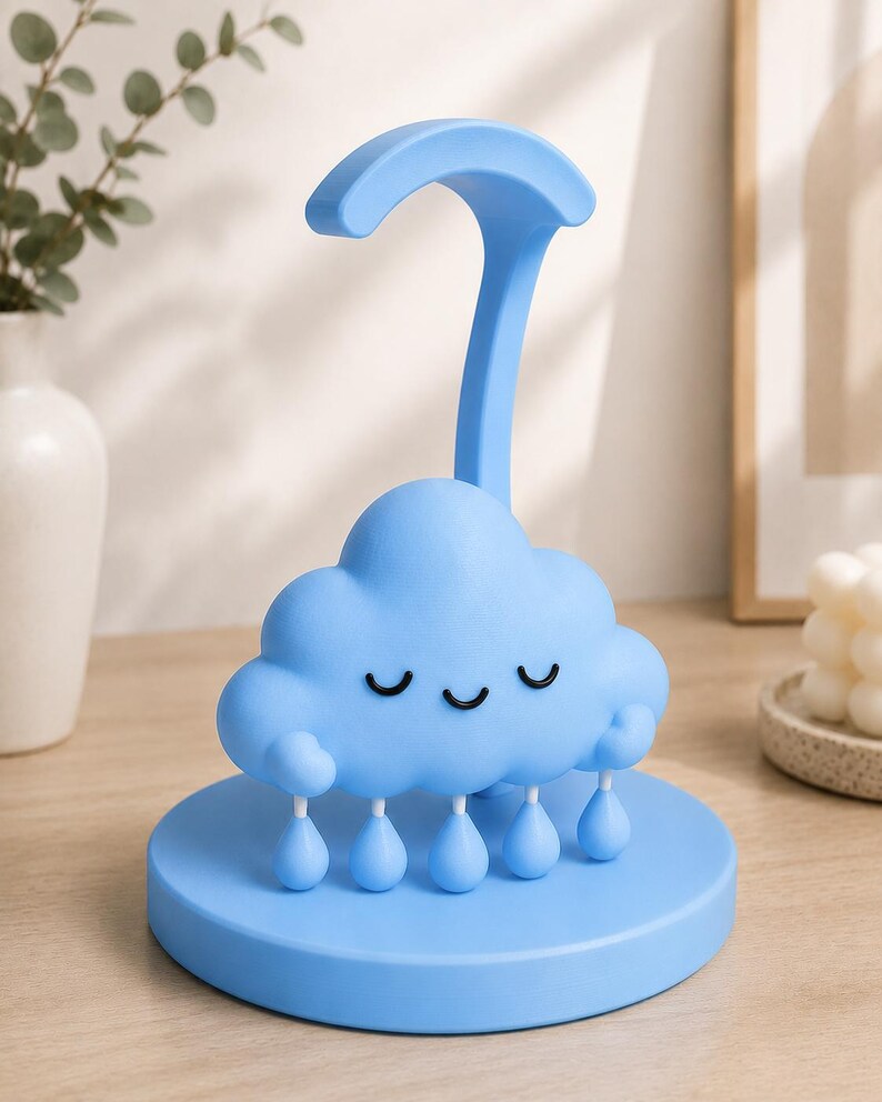 May include: A light blue cloud-shaped night light with a curved handle. The cloud has a smiling face with closed eyes and teardrop-shaped rain drops hanging below. The night light sits on a round base.