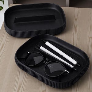 May include: Two black trays with a ribbed design. One tray is empty, the other contains black sunglasses and two white pens. The trays are on a light wood surface.