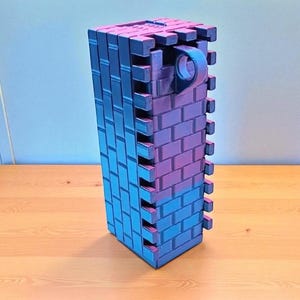 May include: A rectangular, brick-patterned structure in shades of blue and purple. The object has a handle-like feature on one side. The design suggests a decorative or functional item, possibly a container or a building block.