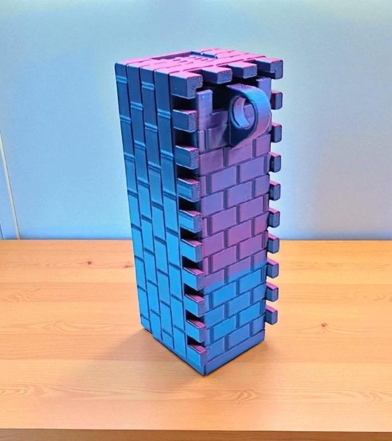 May include: A rectangular, brick-patterned structure in shades of blue and purple. The object has a handle-like feature on one side. The design suggests a decorative or functional item, possibly a container or a building block.