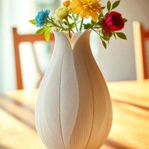 May include: A white, textured vase with a tulip-like design, holding a vibrant bouquet of colorful flowers. The arrangement includes blue, yellow, and red blooms with green foliage. The vase sits on a wooden table.
