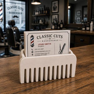 Puede incluir: Un tarjetero de madera con forma de peine sobre una superficie de madera. El soporte contiene una tarjeta de visita con el texto "CLASSIC CUTS BARBERSHOP" e informaci&oacute;n de contacto de John Smith, Barber. El fondo muestra un interior de barber&iacute;a.