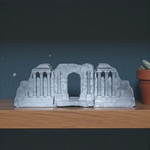 May include: A gray, architectural bookend set. The design features a classical ruin with columns and an arched doorway. The bookends are made of a solid material and are displayed on a wooden shelf.
