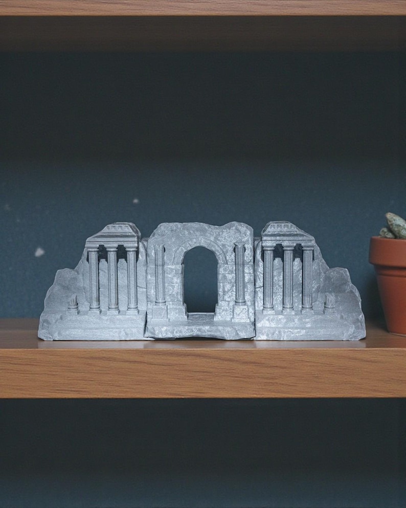 May include: A gray, architectural bookend set. The design features a classical ruin with columns and an arched doorway. The bookends are made of a solid material and are displayed on a wooden shelf.