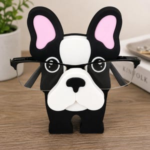 May include: A black and white bulldog-shaped glasses holder with pink inner ears. The holder is designed to hold a pair of eyeglasses. The glasses are black-framed and rest on the bulldog's nose.