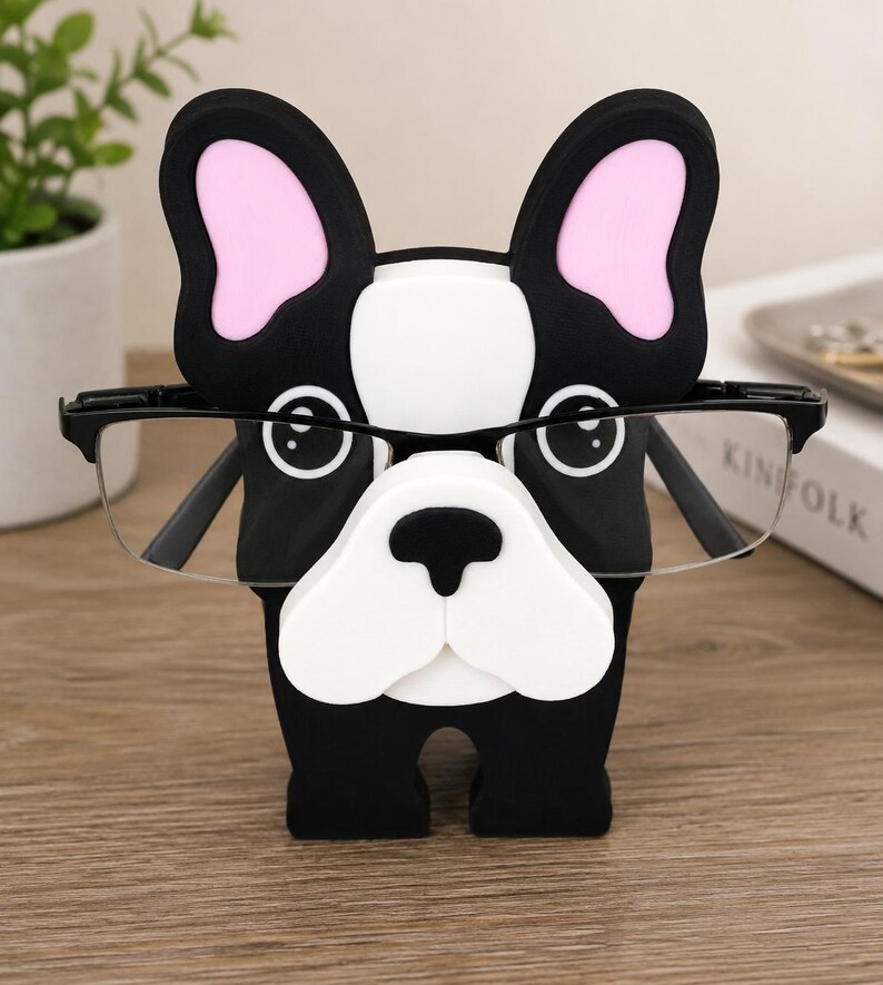 May include: A black and white bulldog-shaped glasses holder with pink inner ears. The holder is designed to hold a pair of eyeglasses. The glasses are black-framed and rest on the bulldog's nose.