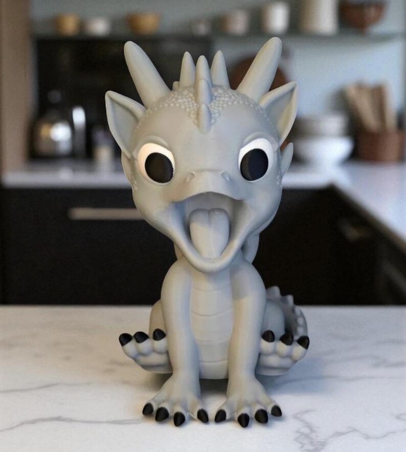May include: A light gray dragon figurine with large black eyes and an open mouth, revealing a pink tongue. The dragon has spiky features on its head and black claws. It is sitting on a white marble surface.