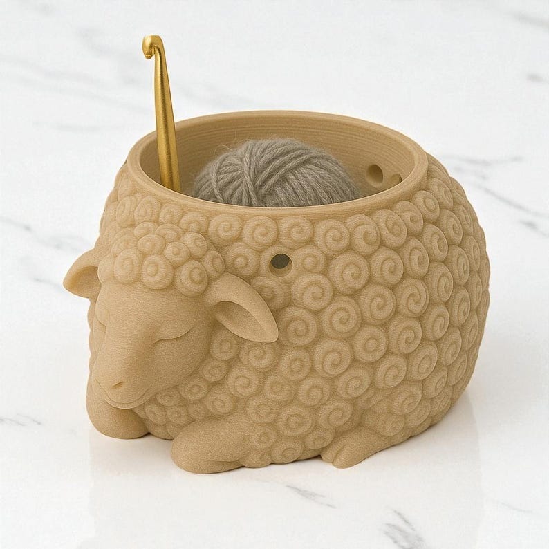 K&ouml;nnte beinhalten: Eine beige schaf&ouml;rmige Garnschale mit einem goldenen H&auml;kelhaken. Die Schale enth&auml;lt einen Kn&auml;uel grauer Wolle. Das Schaf hat ein detailliertes, strukturiertes Wollmuster und liegt auf der Seite.
