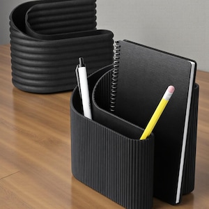 Op de afbeelding: Zwarte bureau-organizer met een geribbelde textuur, met een witte pen, een gele potlood en een zwart notitieboek. Een andere vergelijkbare organizer staat op de achtergrond. De items staan op een houten oppervlak.