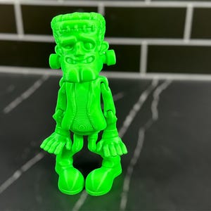 May include: A green plastic figurine of Frankenstein's monster. The figurine is articulated at the shoulders, elbows, wrists, hips, knees, and ankles. The figurine has a detailed face with a stitched scar across the forehead.