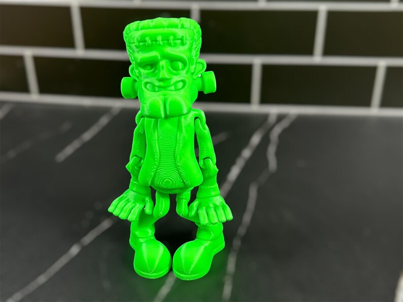 May include: A green plastic figurine of Frankenstein's monster. The figurine is articulated at the shoulders, elbows, wrists, hips, knees, and ankles. The figurine has a detailed face with a stitched scar across the forehead.