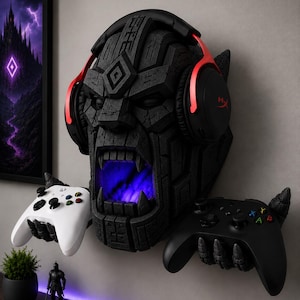 May include: A black, textured, stone-like wall mount shaped as a monstrous head with an open mouth glowing with blue light. It wears black and red over-ear headphones and features two clawed hands extending from the sides to hold a white and a black game controller.