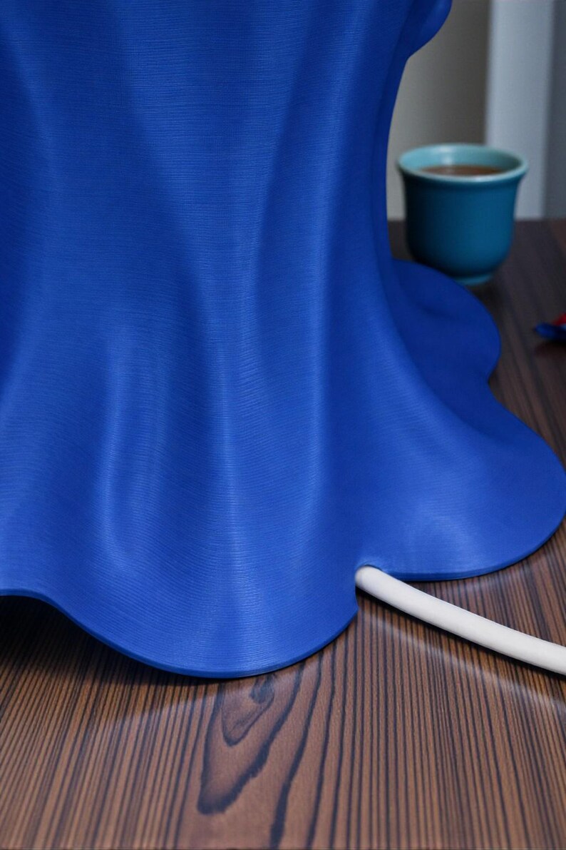 May include: A vibrant blue, petal-shaped object with a white, cylindrical element extending from its base. The object sits on a wooden surface, with a teal cup visible in the background. The object's form suggests a decorative or functional item.