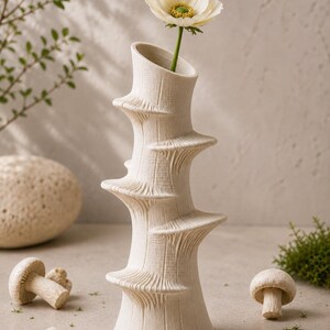 May include: A cream-colored, textured ceramic vase with a unique spiral design, holding a single white flower with a yellow center. The vase is surrounded by decorative mushroom figurines, creating a natural, earthy aesthetic.