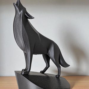 May include: A black, geometric-style wolf sculpture in a howling pose. The wolf stands on a dark, angular base. The sculpture is made of a solid, matte black material and is displayed on a wooden surface.