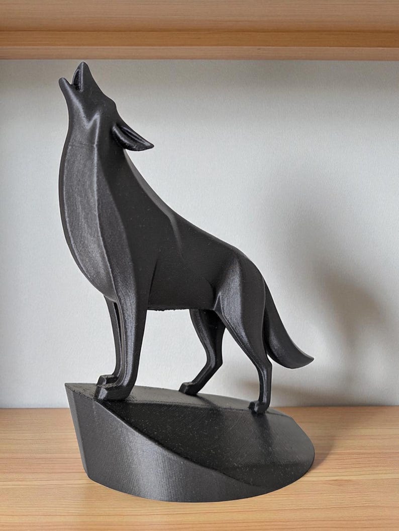 May include: A black, geometric-style wolf sculpture in a howling pose. The wolf stands on a dark, angular base. The sculpture is made of a solid, matte black material and is displayed on a wooden surface.