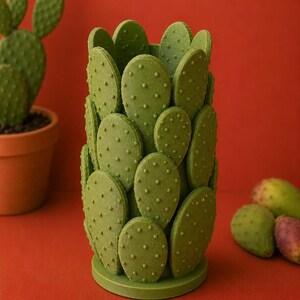 May include: A green cactus-shaped decorative item with a textured surface and small, round bumps. The item is set against a red background, with a potted cactus and several prickly pears visible.