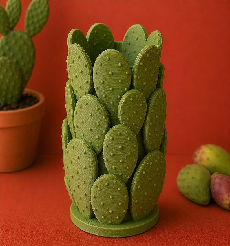 May include: A green cactus-shaped decorative item with a textured surface and small, round bumps. The item is set against a red background, with a potted cactus and several prickly pears visible.
