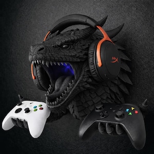 Puede incluir: Una escultura de cabeza de dragón negro con auriculares naranjas y negros y dos mandos de juego. La boca del dragón está abierta, revelando afilados dientes y un brillo azul. Un mando blanco y otro negro se sostienen en las manos del dragón.