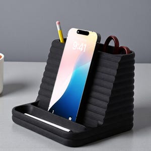 May include: A black desk organizer with a built-in phone stand. The organizer has a ribbed texture and holds a smartphone, a pencil, and scissors. The phone displays a colorful wallpaper. A small tray is at the base.