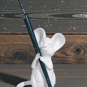 May include: A white mouse figurine holding a dark blue paintbrush. The mouse has large ears and a curved tail. The figurine is set against a wooden background.