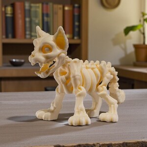 Puede incluir: Figura de esqueleto de gato impresa en 3D, de color crema. La figura tiene una estructura esquel&eacute;tica detallada con costillas, columna vertebral y cr&aacute;neo visibles. Los ojos y las orejas tienen un tono amarillo-anaranjado. El gato est&aacute; en posici&oacute;n de caminar.