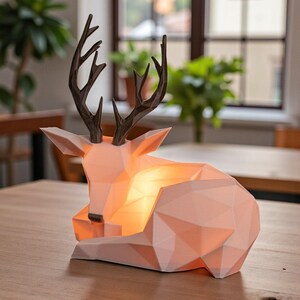 Puede incluir: L&aacute;mpara con forma de ciervo geom&eacute;trico rosa claro con cuernos marr&oacute;n oscuro. La l&aacute;mpara est&aacute; sobre una mesa de madera, con una luz c&aacute;lida que emana del interior. Dise&ntilde;o moderno y minimalista.