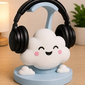 May include: A whimsical headphone stand in the shape of a smiling cloud. The stand is light blue with a white cloud figure featuring pink cheeks and a black smiling mouth. Black headphones rest on the stand.