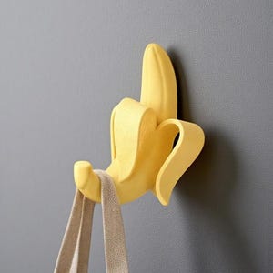 May include: A yellow banana-shaped wall hook with a beige tote bag hanging from it. The hook is mounted on a grey wall. The banana peel is partially open, revealing the hook. The tote bag is made of a light-coloured fabric.