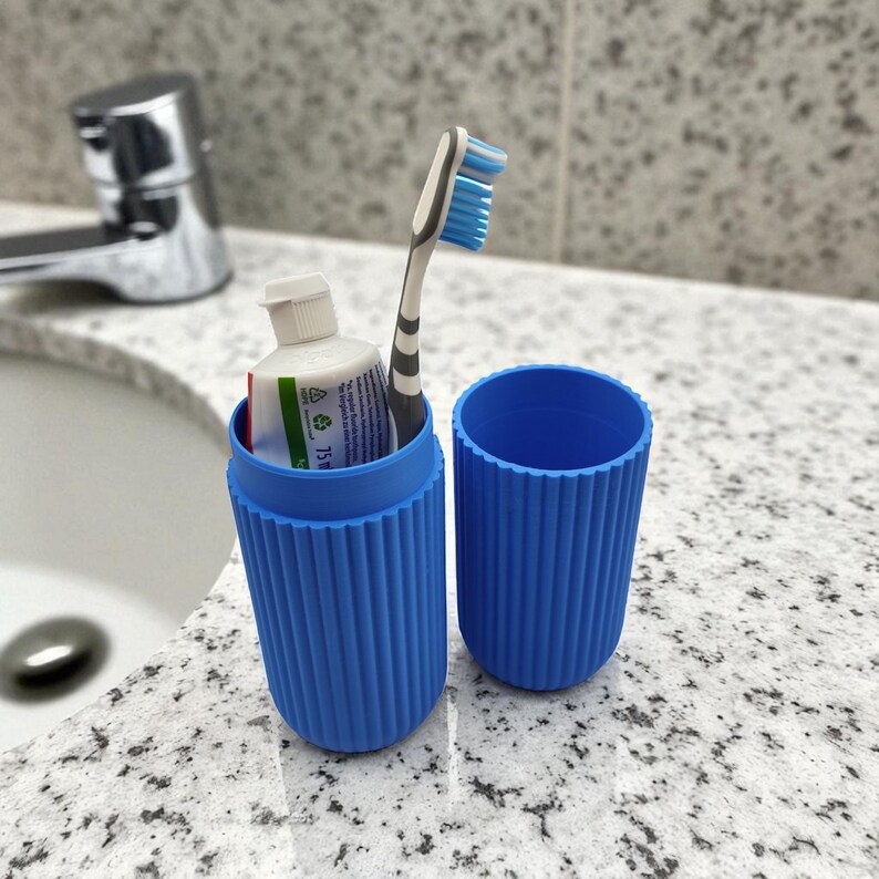 Pode incluir: Um porta-escova de dentes de viagem azul canelado com uma escova de dentes e pasta de dentes dentro. H&aacute; outro recipiente vazio ao lado. Os itens est&atilde;o em uma bancada salpicada em um banheiro.