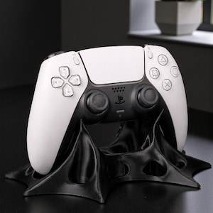 May include: A white and black PlayStation 5 controller resting on a black stand. The controller has a white top and black bottom with various button symbols. The stand has a unique, flowing design.