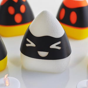 May include: A set of five white candy corn shaped candle holders with black and orange Halloween themed designs. The candle holders have a smiling face, a jack-o-lantern face, and two different polka dot designs.