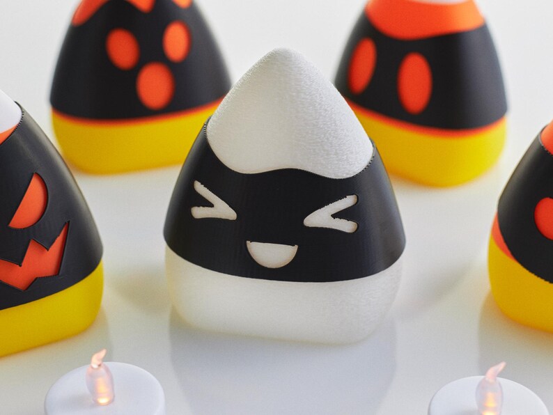 May include: A set of five white candy corn shaped candle holders with black and orange Halloween themed designs. The candle holders have a smiling face, a jack-o-lantern face, and two different polka dot designs.