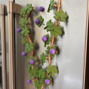 May include: A decorative grape vine with green leaves and purple grapes, attached to a refrigerator. The vine is made of wood and is arranged in a curved shape. The grapes are round and evenly spaced.