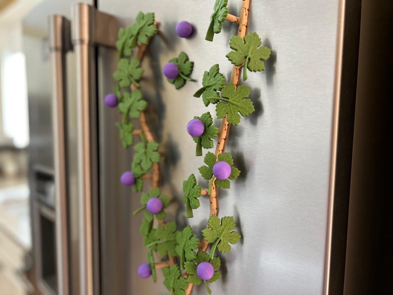May include: A decorative grape vine with green leaves and purple grapes, attached to a refrigerator. The vine is made of wood and is arranged in a curved shape. The grapes are round and evenly spaced.