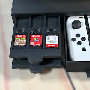 May include: A black plastic storage case with a drawer that holds three Nintendo Switch game cartridges. The drawer is open and the cartridges are visible. The case also has a compartment for two white Nintendo Switch Joy-Con controllers.
