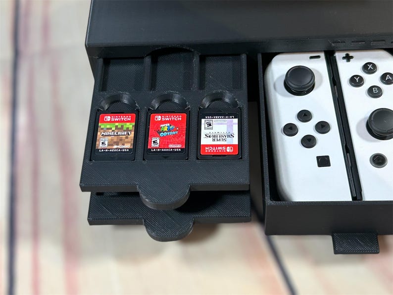 May include: A black plastic storage case with a drawer that holds three Nintendo Switch game cartridges. The drawer is open and the cartridges are visible. The case also has a compartment for two white Nintendo Switch Joy-Con controllers.