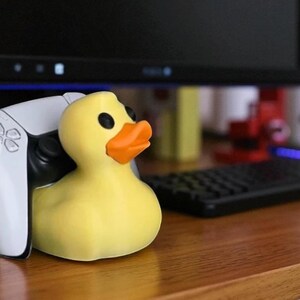 May include: A yellow rubber duck with black eyes and an orange beak. The duck is positioned next to a white game controller. The background includes a computer monitor and a black keyboard, suggesting a gaming setup.