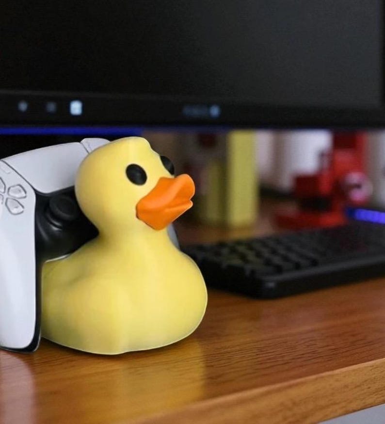 May include: A yellow rubber duck with black eyes and an orange beak. The duck is positioned next to a white game controller. The background includes a computer monitor and a black keyboard, suggesting a gaming setup.