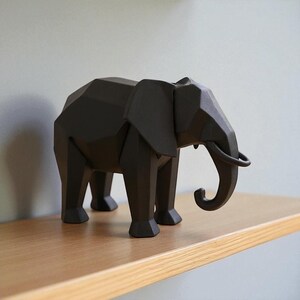 Op de afbeelding: Een geometrisch, zwart olifantenbeeld met een slurf, oren en slagtanden. De olifant staat op een houten plank. Het beeld heeft een modern, hoekig ontwerp en is een decoratief item.
