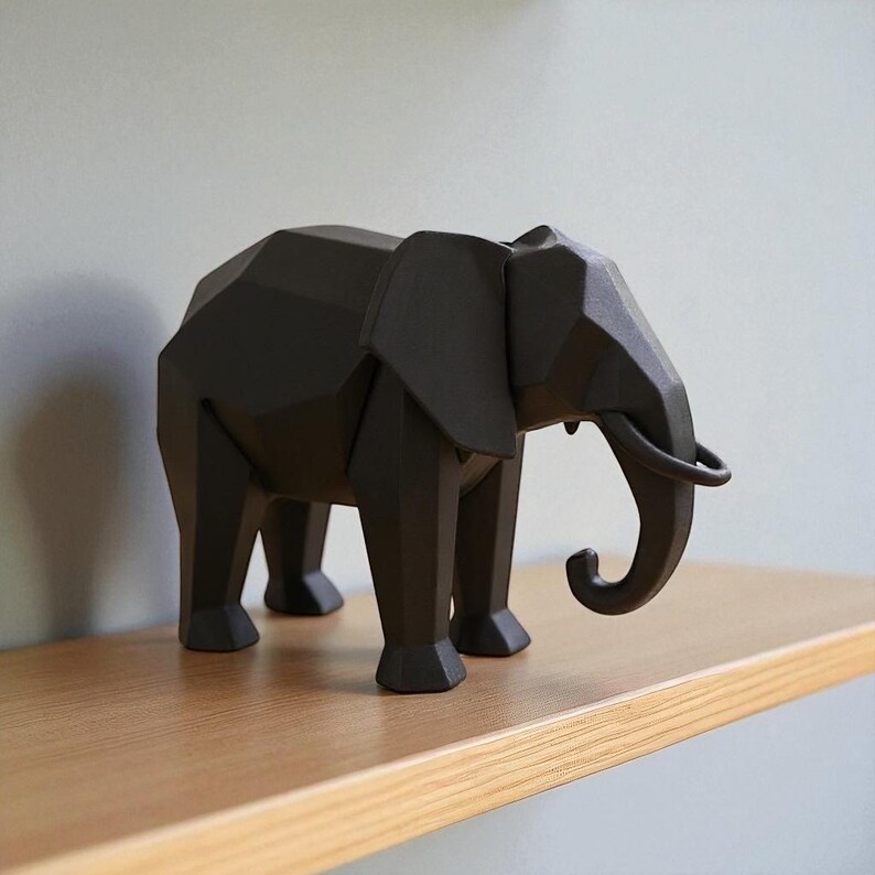 Op de afbeelding: Een geometrisch, zwart olifantenbeeld met een slurf, oren en slagtanden. De olifant staat op een houten plank. Het beeld heeft een modern, hoekig ontwerp en is een decoratief item.