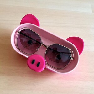 Op de afbeelding: Een roze varkensvormige tray met een zonnebril. De tray heeft een roze snuit met zwarte neusgaten en twee roze oren. De zonnebril heeft een gouden montuur en donkere glazen.