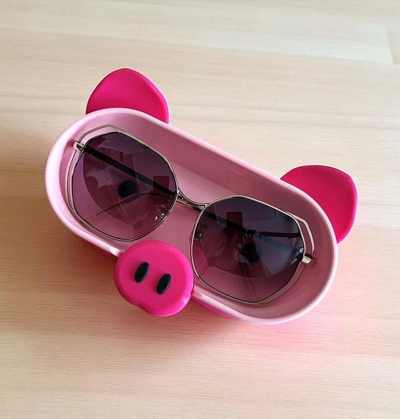 Op de afbeelding: Een roze varkensvormige tray met een zonnebril. De tray heeft een roze snuit met zwarte neusgaten en twee roze oren. De zonnebril heeft een gouden montuur en donkere glazen.