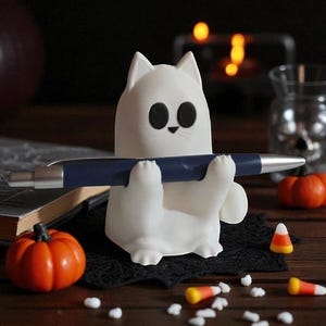 May include: A white cat-shaped pen holder with black eyes and a small nose, holding a blue pen. The pen holder is on a black lace doily, with small orange pumpkins and candy corn scattered around. A Halloween-themed desk accessory.