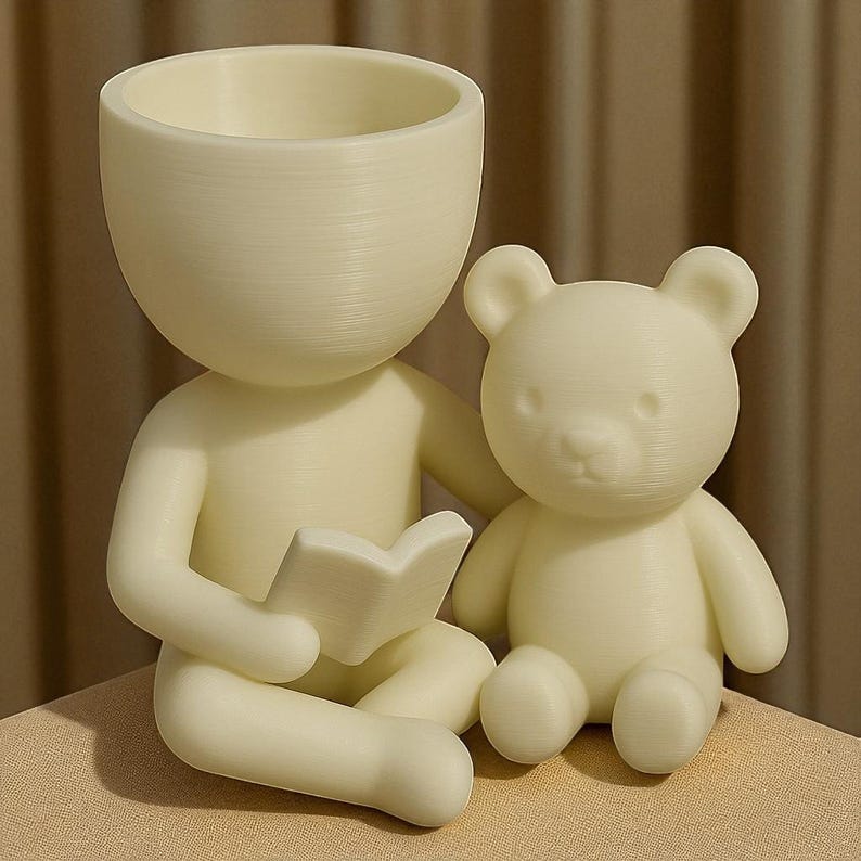 May include: A cream-coloured planter shaped like a person reading a book, with a teddy bear sitting beside them. The person-shaped planter has a large head with a hollowed-out top for planting. The teddy bear is small and sitting upright.