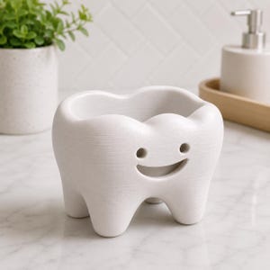 Może przedstawiać: Biały ceramiczny pojemnik w kształcie zęba z uśmiechniętą twarzą. Pojemnik ma wydrążony wierzch i jest podparty trzema małymi n&oacute;żkami. Tło obejmuje roślinę w białej doniczce i dozownik mydła.