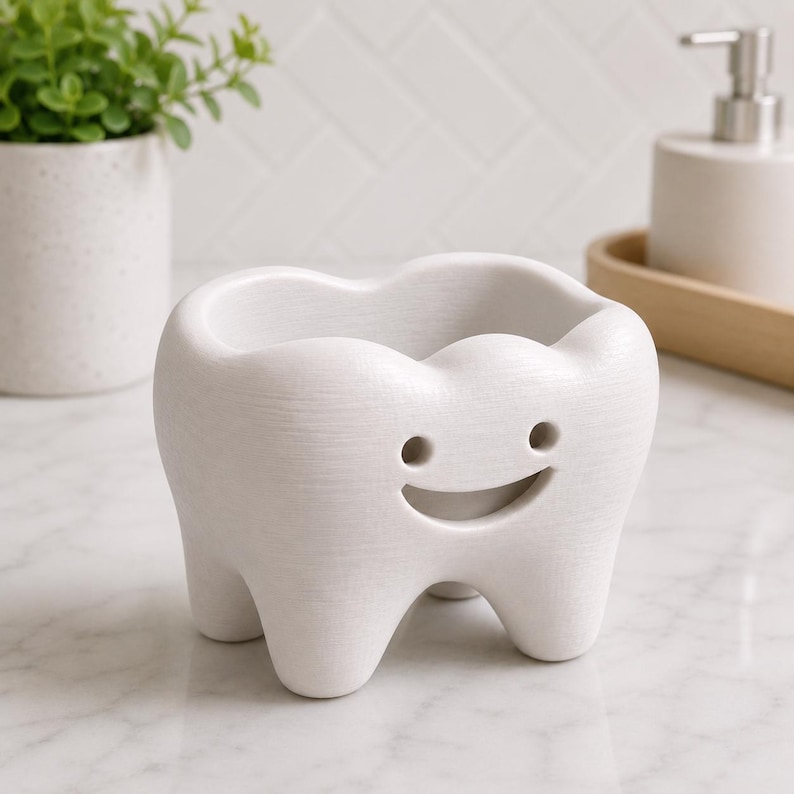 Może przedstawiać: Biały ceramiczny pojemnik w kształcie zęba z uśmiechniętą twarzą. Pojemnik ma wydrążony wierzch i jest podparty trzema małymi n&oacute;żkami. Tło obejmuje roślinę w białej doniczce i dozownik mydła.