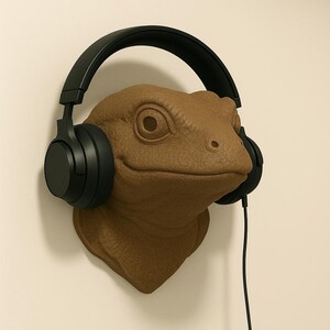 May include: A brown lizard head wall mount with black headphones. The lizard has detailed skin texture and is mounted on a neutral-coloured wall. The headphones have a black cable.