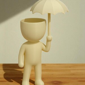 May include: A cream-colored figurine of a person holding an umbrella. The figure has a large, open head, functioning as a planter. The umbrella is also cream-colored, providing a whimsical design. The figurine stands on a wooden surface.